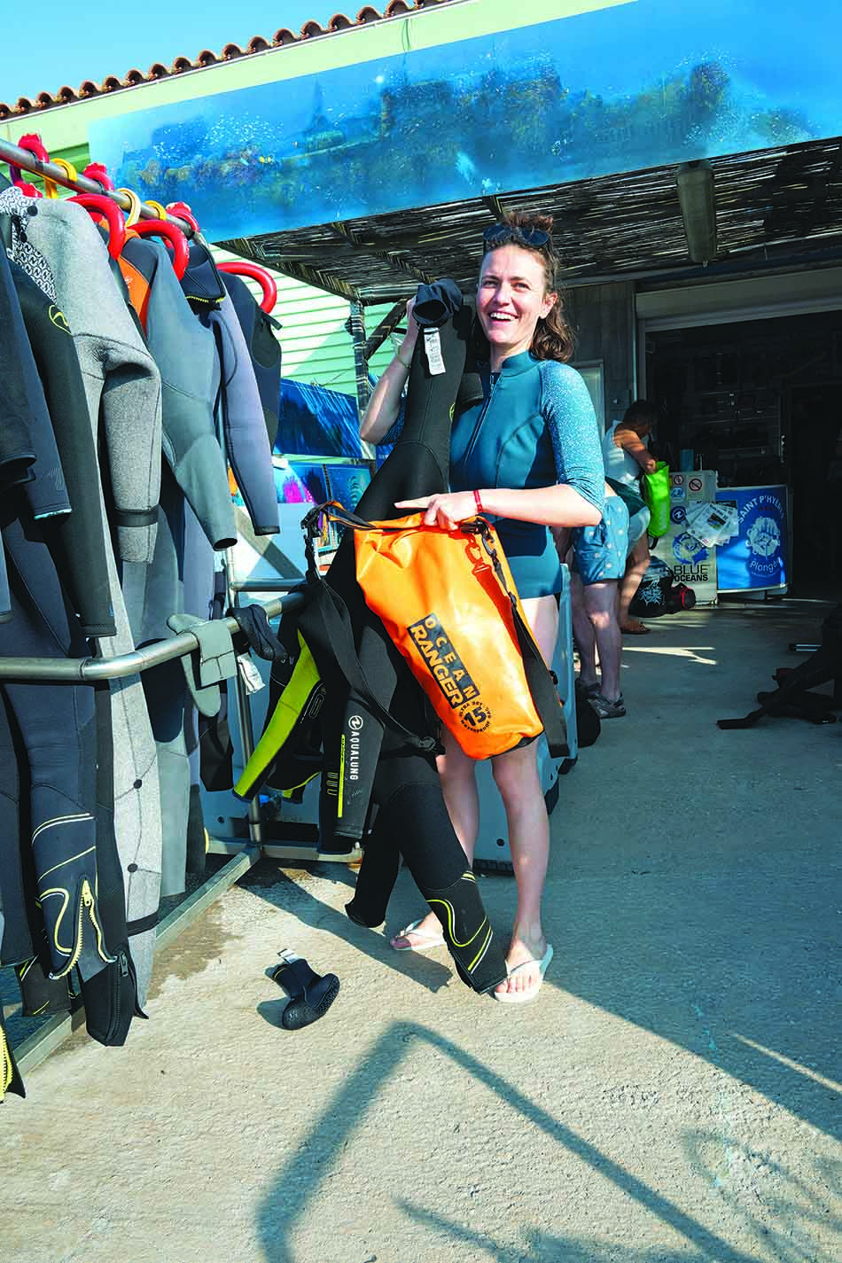 A smiling diver chooses her diving equipment near a busy dive center.