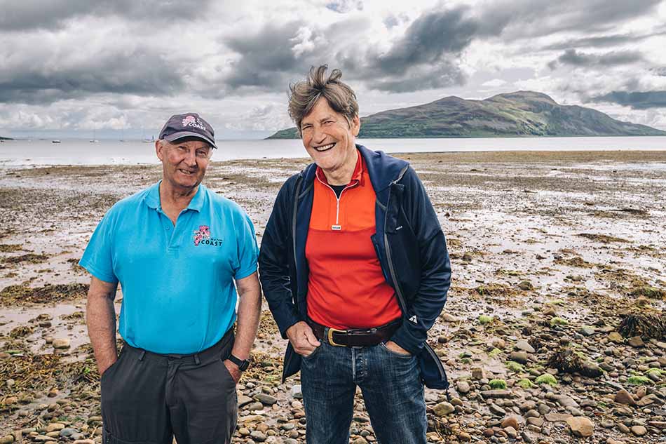 Two diving enthusiasts pose on the coast, with a marine landscape in the background.